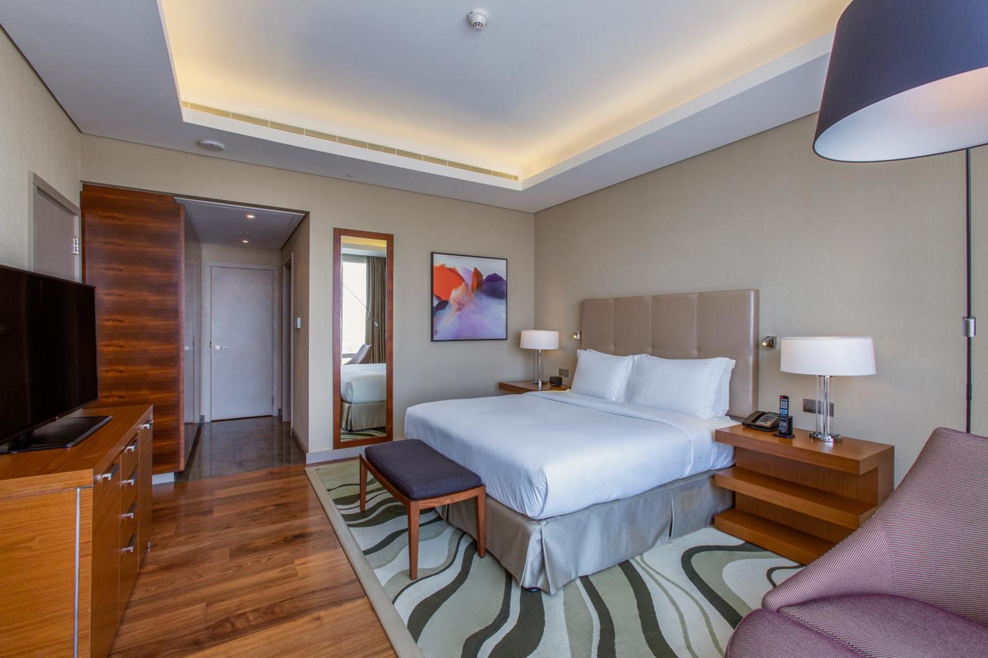 Hilton Riyadh Hotel & Residences Exteriör bild The photo shows a modern hotel room with a comfortable and stylish design. It features a large bed with white linens and a headboard, flanked by two bedside lamps. There is a small bench at the foot of the bed and a cozy chair in the corner. A flat-s