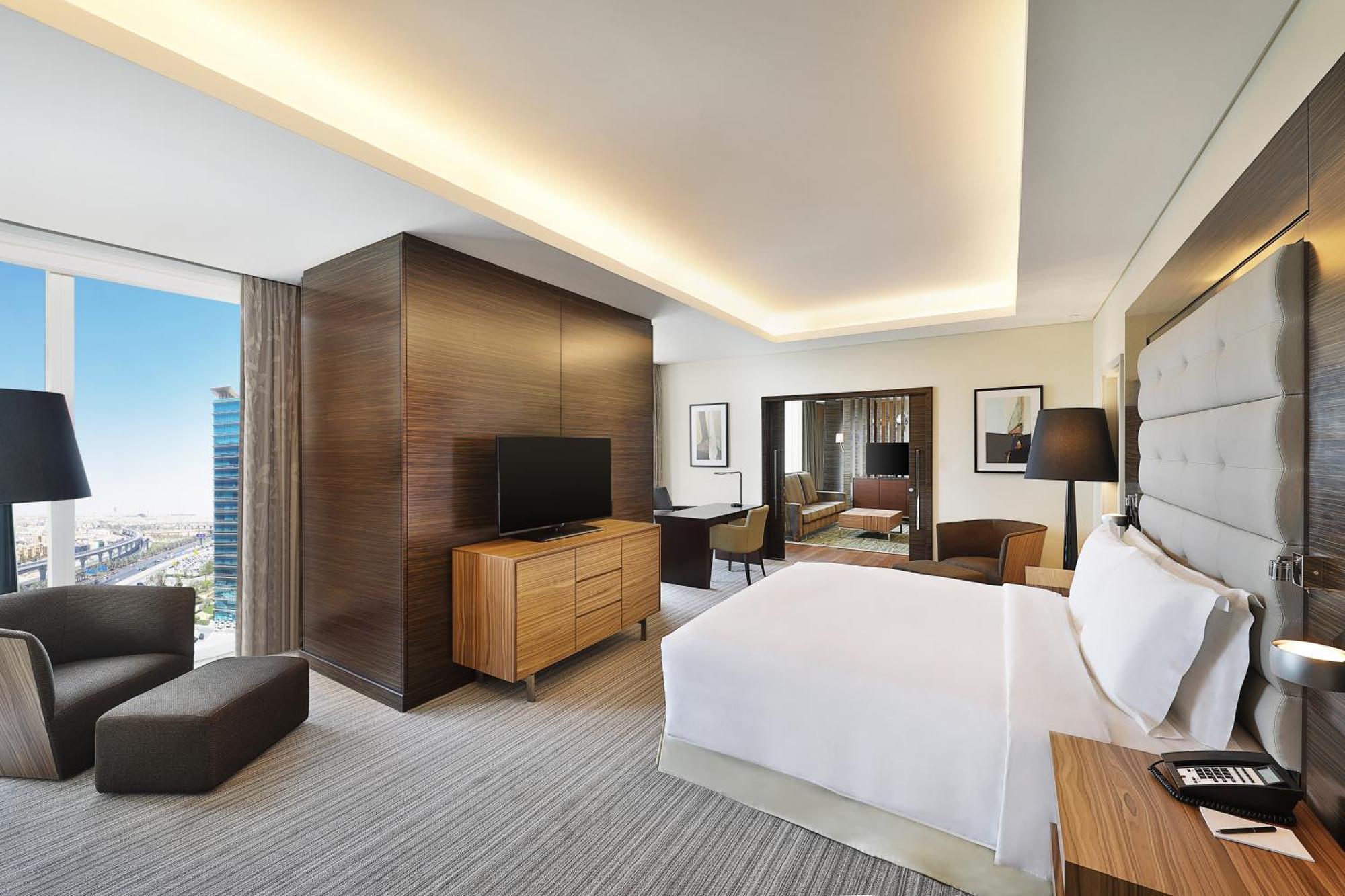 Hilton Riyadh Hotel & Residences Exteriör bild The photo shows a modern hotel room. It features a neatly made bed with white linens, a wooden nightstand beside it, and a cozy armchair. The room has a large window that provides a view of the outside, likely showcasing a cityscape. A flat-screen TV