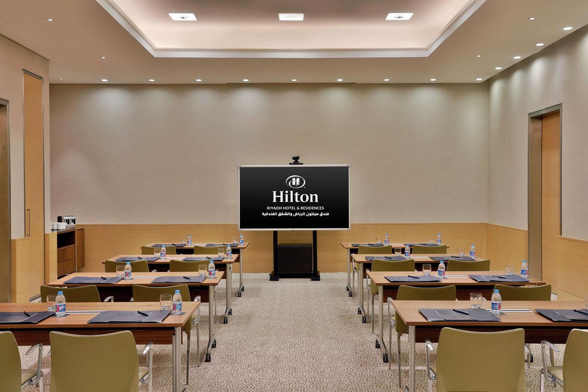 Hilton Riyadh Hotel & Residences Exteriör bild The photo shows a conference or meeting room set up for a presentation. There are several tables arranged in a classroom style, each equipped with chairs. On each table, there are notepads and water bottles. At the front of the room is a large screen
