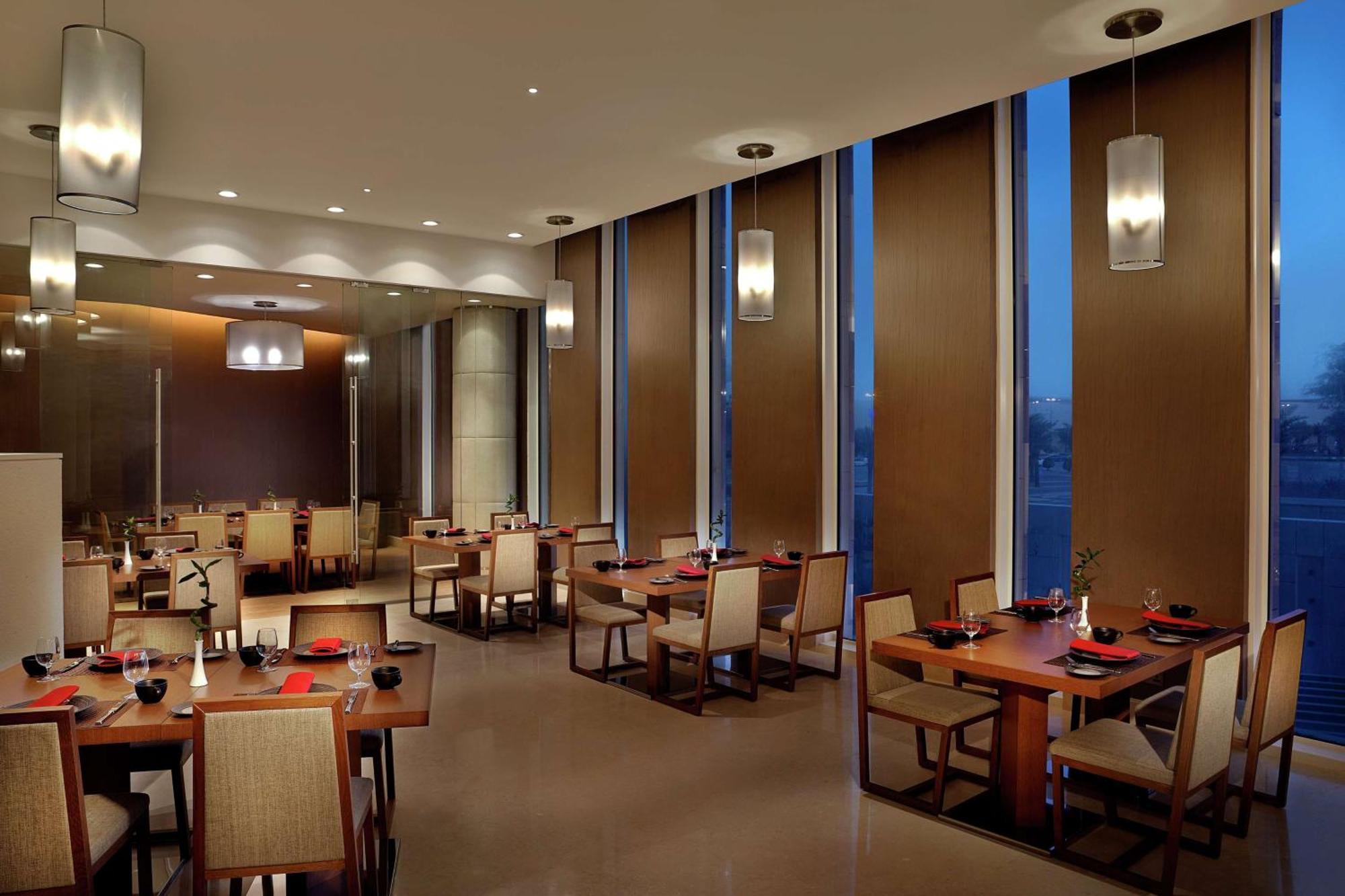Hilton Riyadh Hotel & Residences Exteriör bild The image depicts a modern restaurant interior. It features several dining tables arranged neatly, each set with plates and utensils. The room has large windows with blinds partially drawn, allowing some ambient light to filter in, while evening ligh