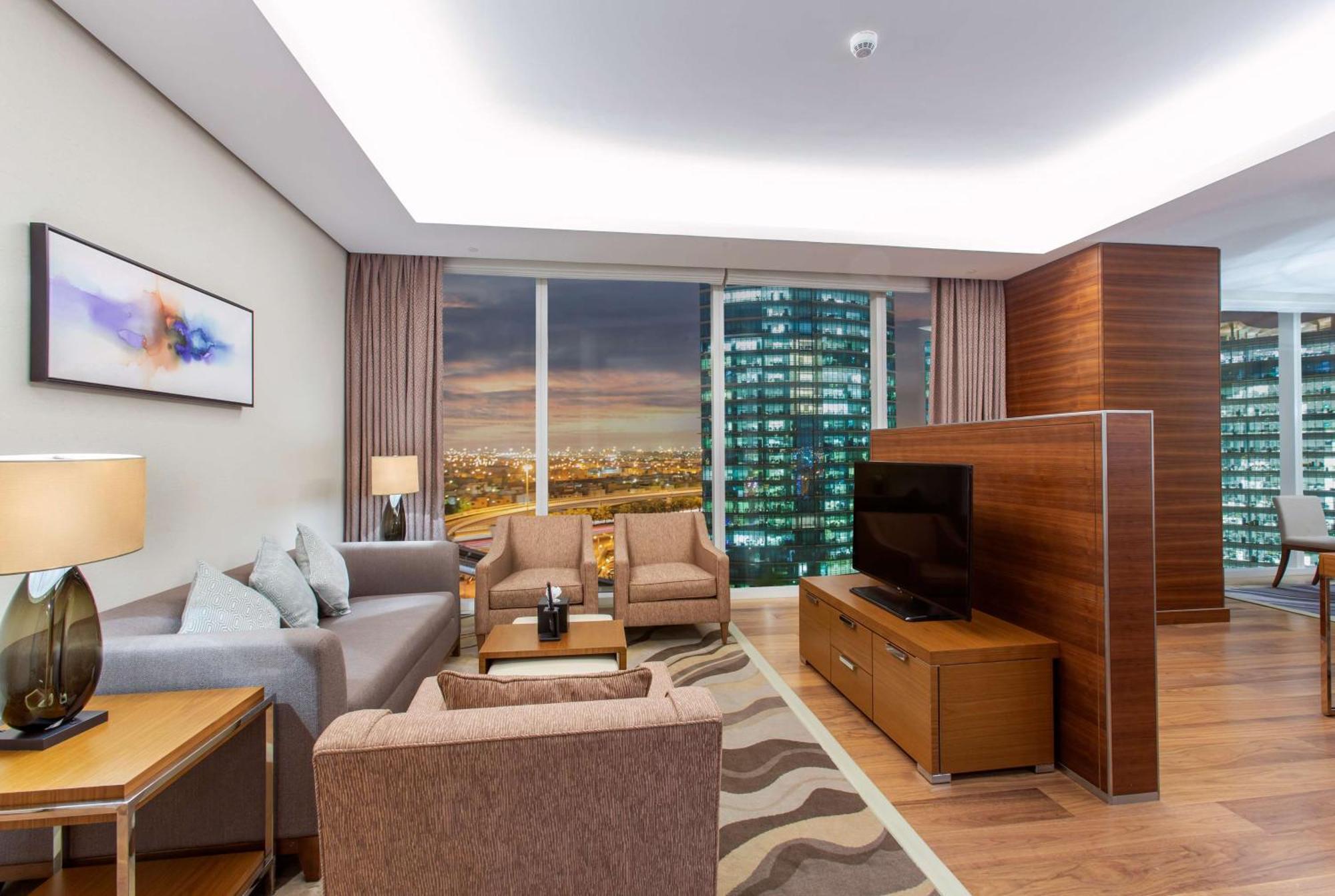 Hilton Riyadh Hotel & Residences Exteriör bild The photo shows a modern living room in a stylish apartment. The space features a comfortable seating arrangement with a light gray sofa and two brown armchairs, complemented by a wooden coffee table. There is a large flat-screen TV mounted on a wood