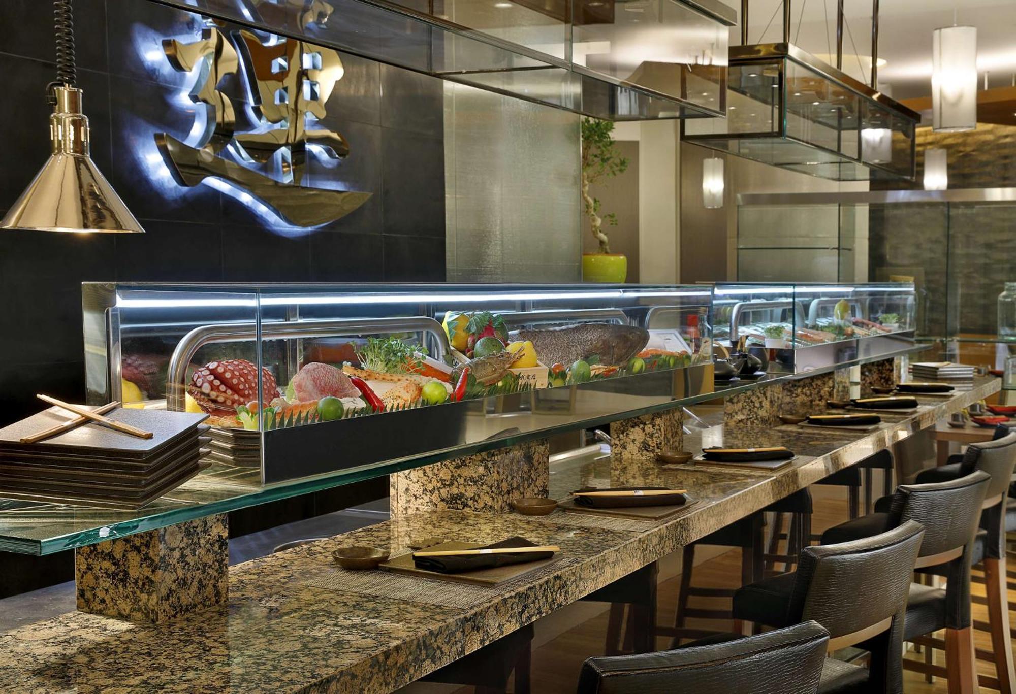 Hilton Riyadh Hotel & Residences Exteriör bild The photo shows an interior view of a sushi restaurant. There is a long, sleek sushi bar with a glass display case filled with various sushi and sashimi offerings. The counter is made of polished stone, and there are several dark chairs arranged neat